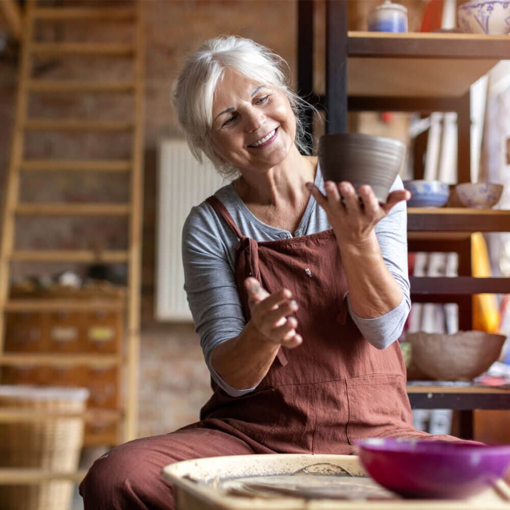Smiling elderly woman in an apron, seated, shaping a clay pot in a pottery studio. - Home Instead