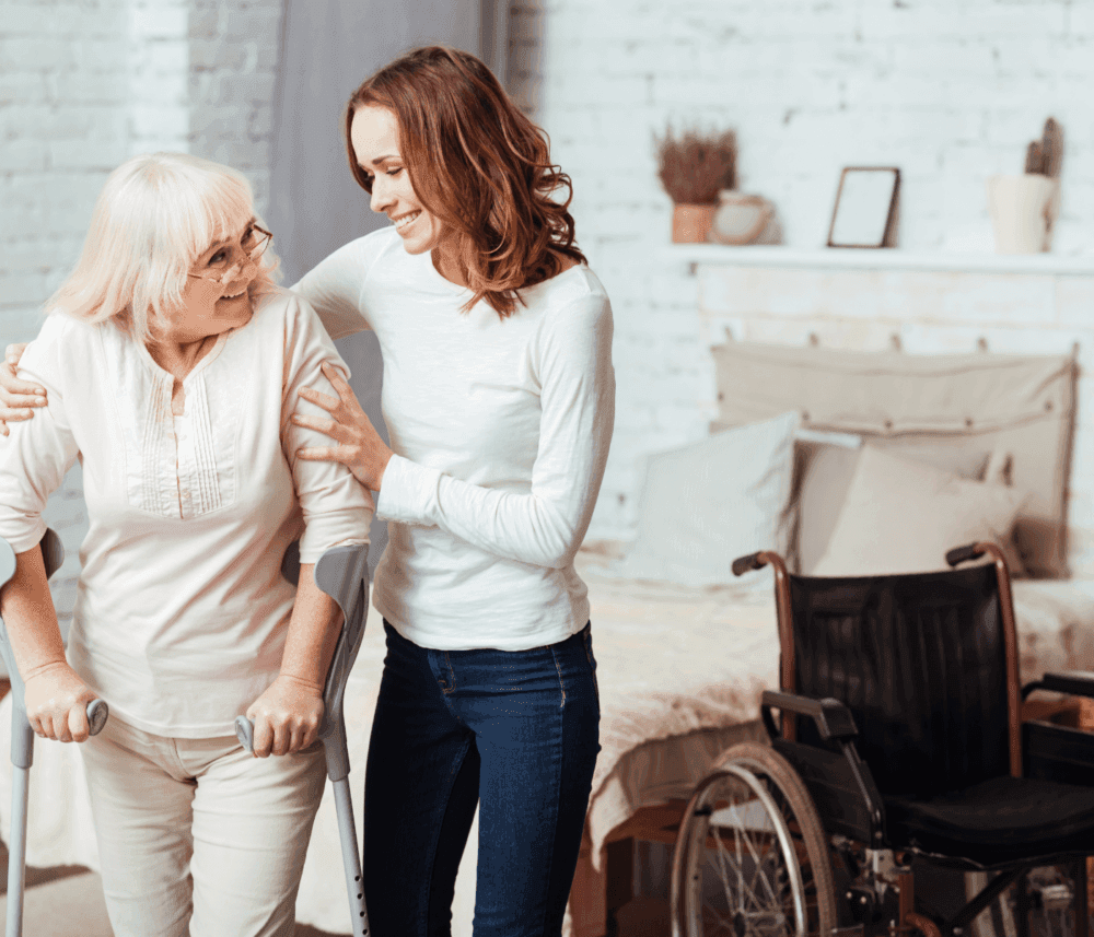 A woman helps an elderly lady using crutches, with a wheelchair in the background, in a cozy, well-lit room. - Home Instead