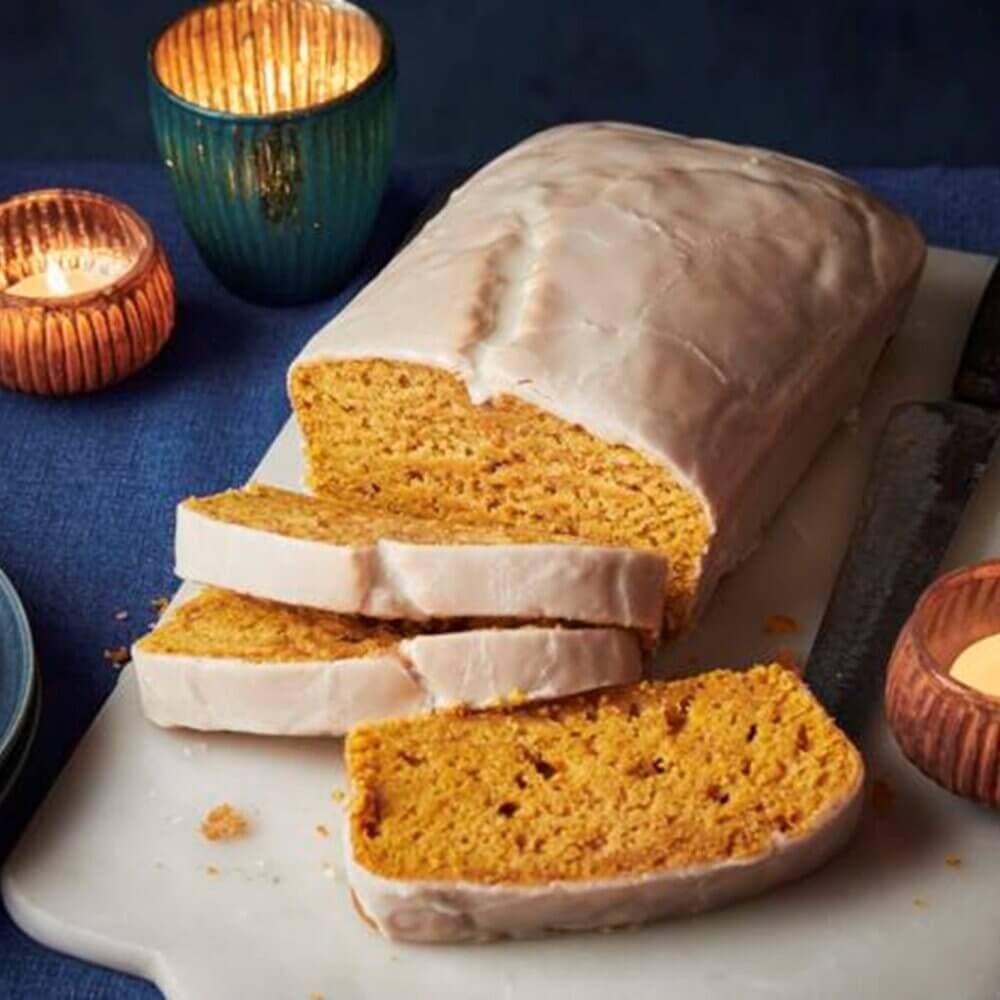 Loaf of iced pumpkin bread partially sliced on a marble board next to a lit candle. - Home Instead