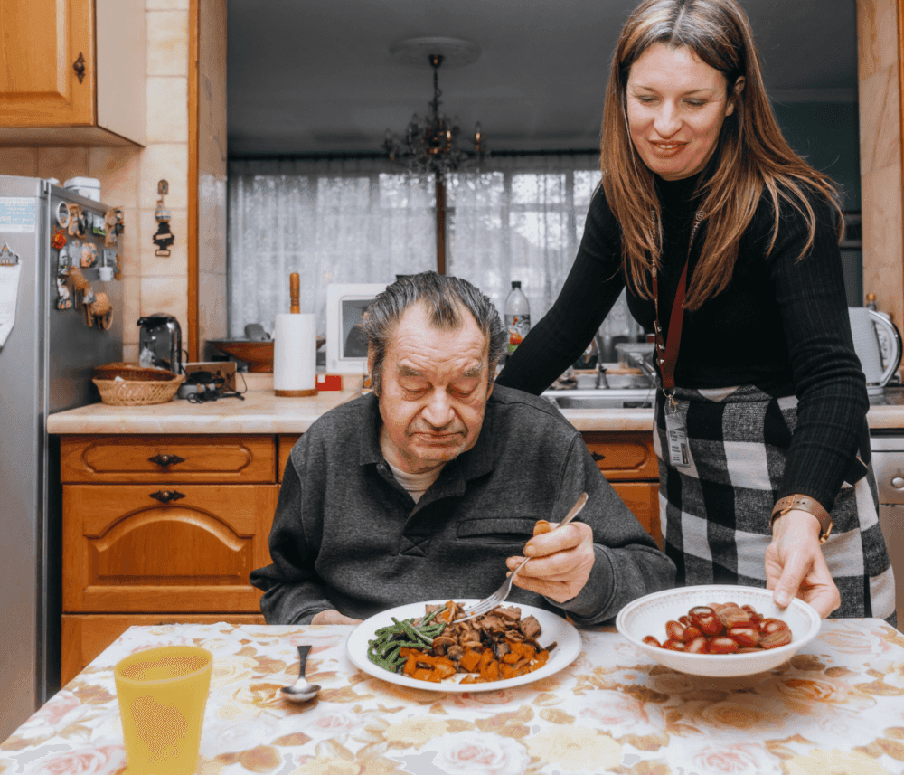 A woman assists an elderly man eating a meal at a kitchen table, handing him a bowl of food. - Home Instead