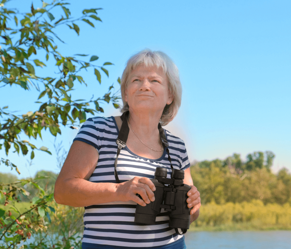 Older woman in a striped shirt holds binoculars, standing outdoors with trees and a clear sky in the background. - Home Instead
