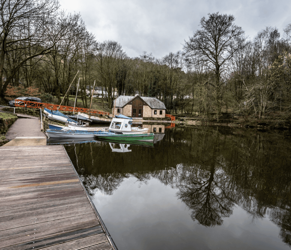Boats docked on a quiet lake beside a wooden pier, with a cabin and dense trees reflected in the calm water. - Home Instead