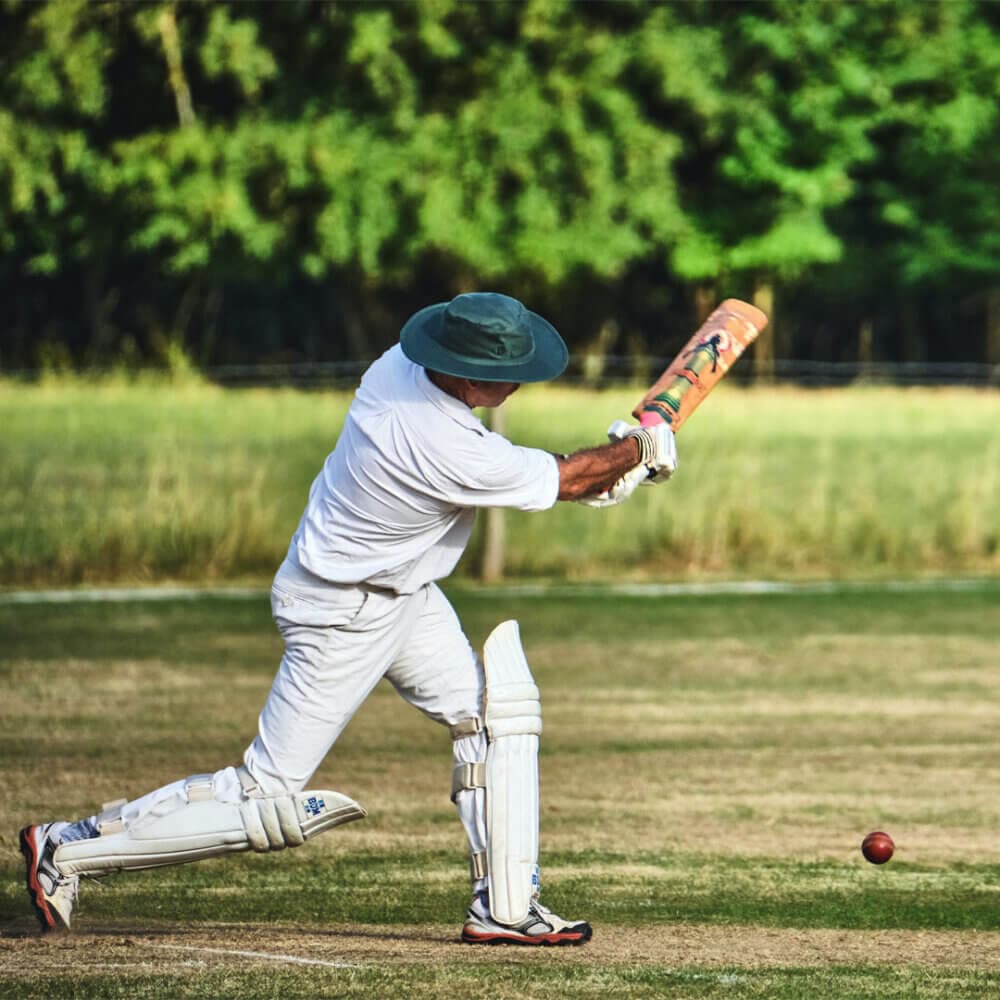 A cricketer in white uniform swings his bat to hit a ball on a grassy field with trees in the background. - Home Instead