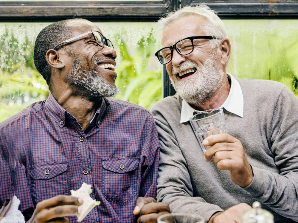 Two elderly men, one wearing a checkered shirt and the other in a gray sweater, laugh together while holding drinks. - Home Instead