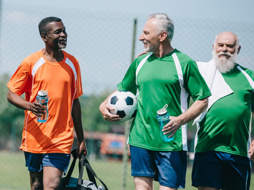 Three smiling men in sports jerseys, one holding a soccer ball, chatting and carrying water bottles on a field. - Home Instead