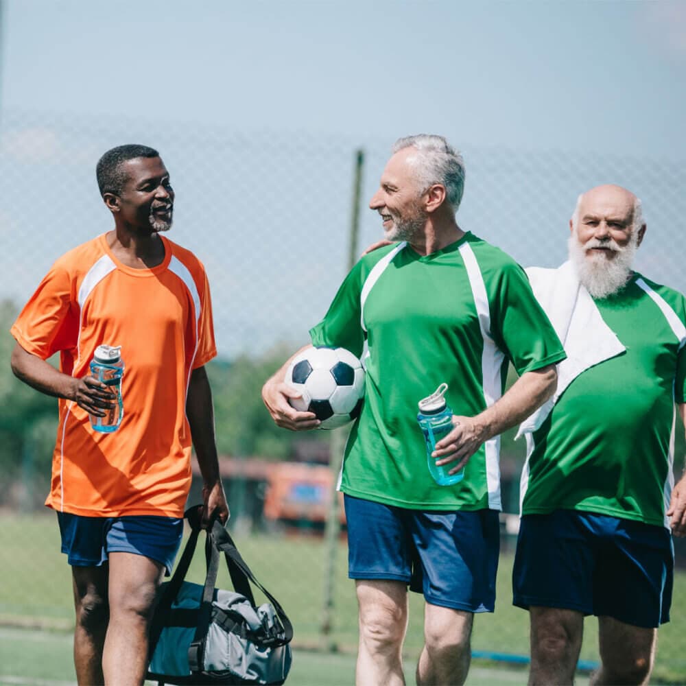 Three senior men in sports attire are smiling and chatting on a soccer field, holding a soccer ball and water bottles. - Home Instead