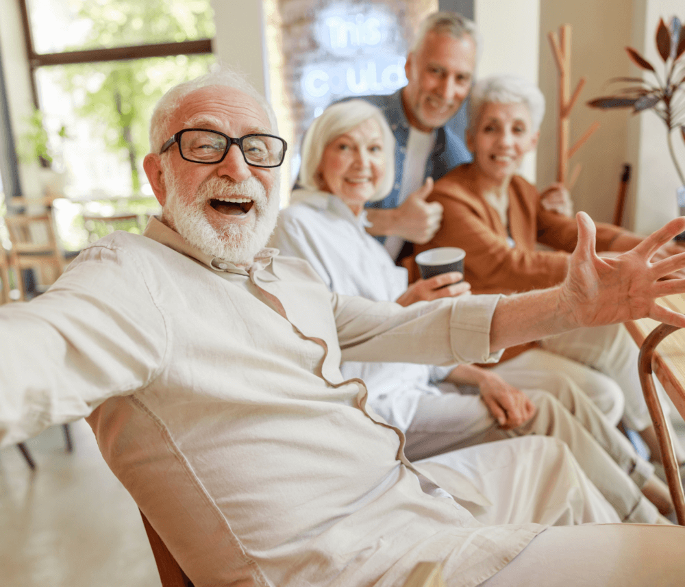 Four cheerful elderly individuals sitting together, smiling and posing with arms outstretched inside a cozy cafe. - Home Instead