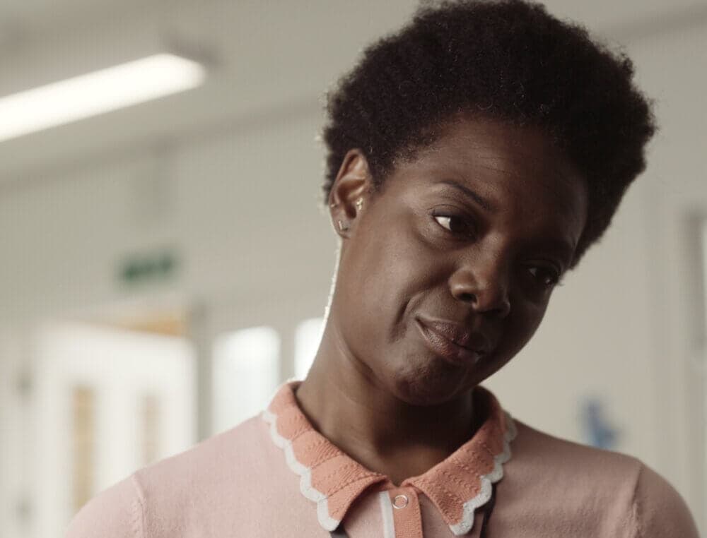 A person with short curly hair looks contemplative, wearing a pink top with a white scalloped collar. - Home Instead