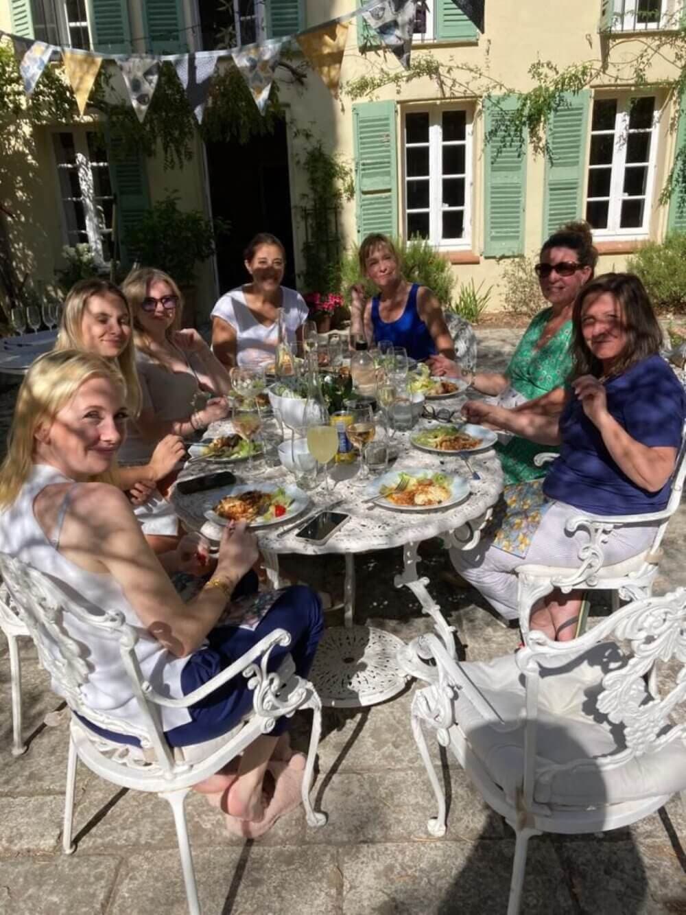 Group of women enjoying a meal together outdoors at a round table, with a house and hanging bunting in the background. - Home Instead