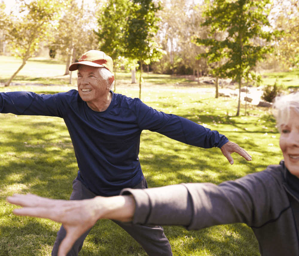 Elderly man and woman smiling and stretching arms during outdoor exercise in a sunny park. - Home Instead