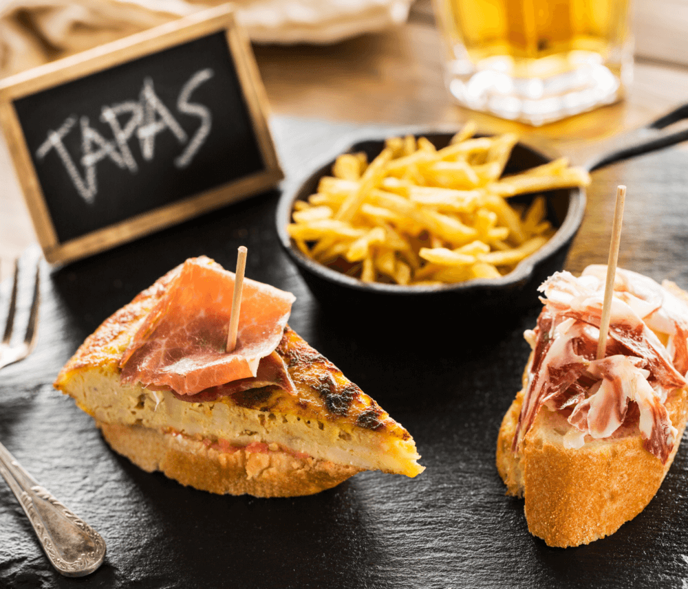 Two tapas dishes with cured ham and omelette on bread, accompanied by a small bowl of fries and a drink in the background. - Home Instead