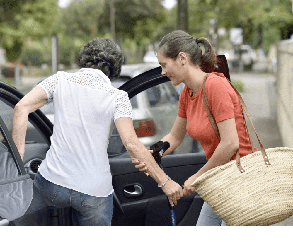 A woman helps an elderly lady with a cane get into a car while holding her arm on a street. - Home Instead