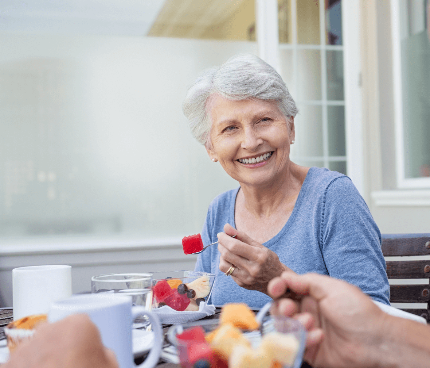 Smiling elderly woman in a blue shirt enjoying a fruit salad at an outdoor table with others. - Home Instead