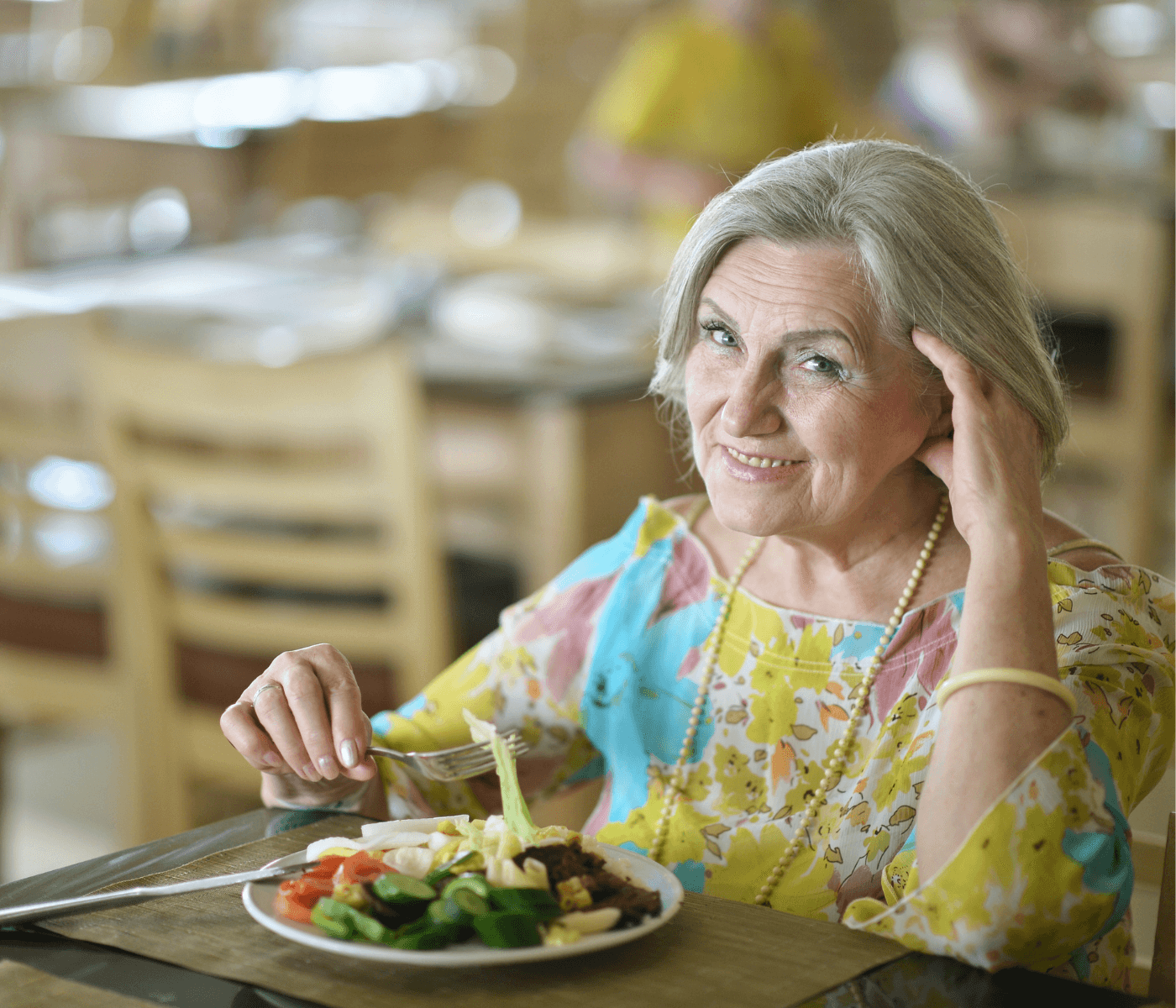 An elderly woman with gray hair smiles while sitting at a table with a meal in a brightly lit dining area. - Home Instead