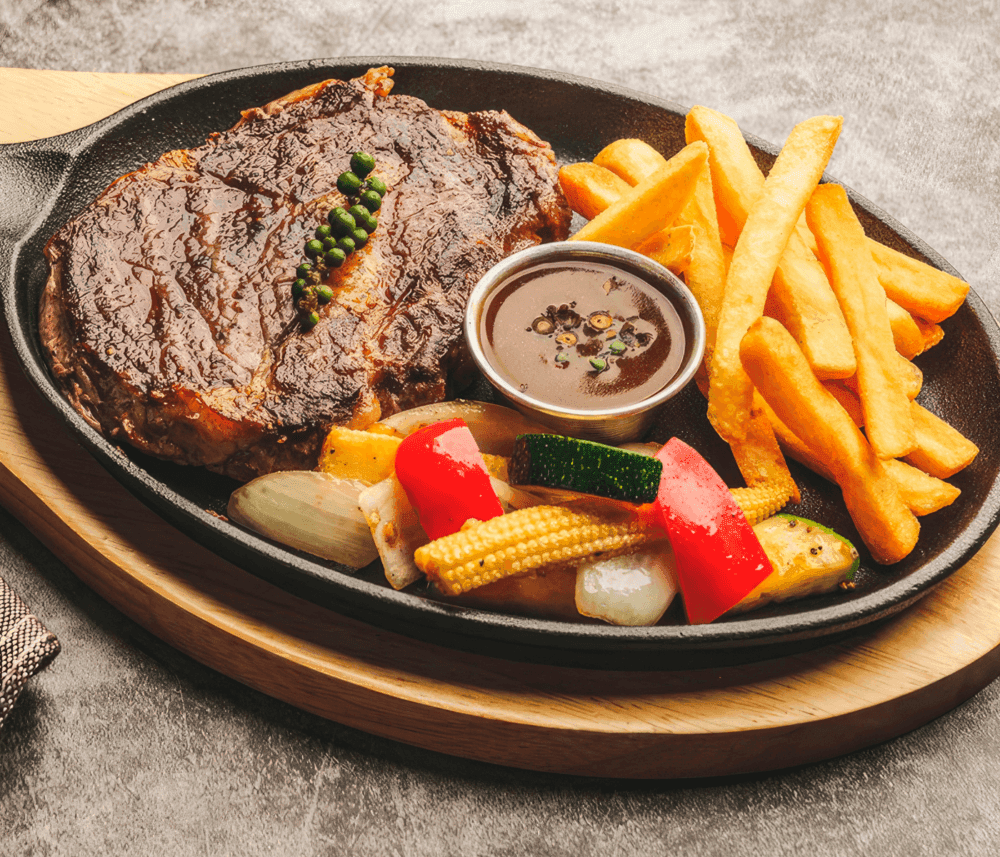 A steak with peppercorns, fries, mixed grilled vegetables, and a small bowl of sauce on a sizzling hot plate. - Home Instead