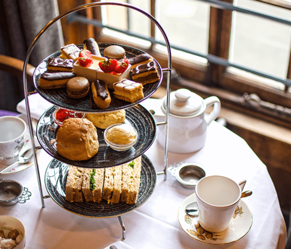 A three-tiered stand with sandwiches, pastries, and desserts next to a teapot, cup, and saucer on a table by a window. - Home Instead