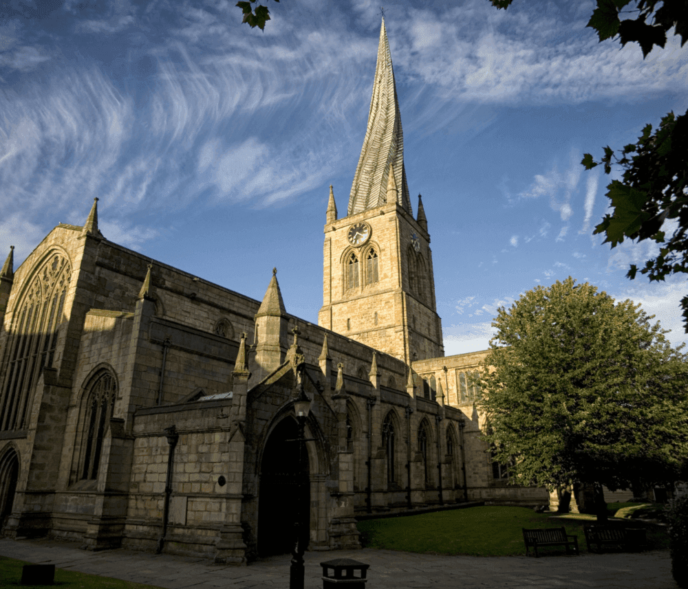 A historic stone church with a tall, twisted spire under a blue sky with wispy clouds, surrounded by trees. - Home Instead