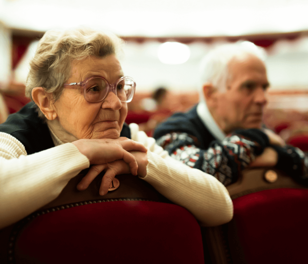 Two elderly people, one in glasses, sit close to each other, leaning forward on red theater seats, gazing ahead. - Home Instead