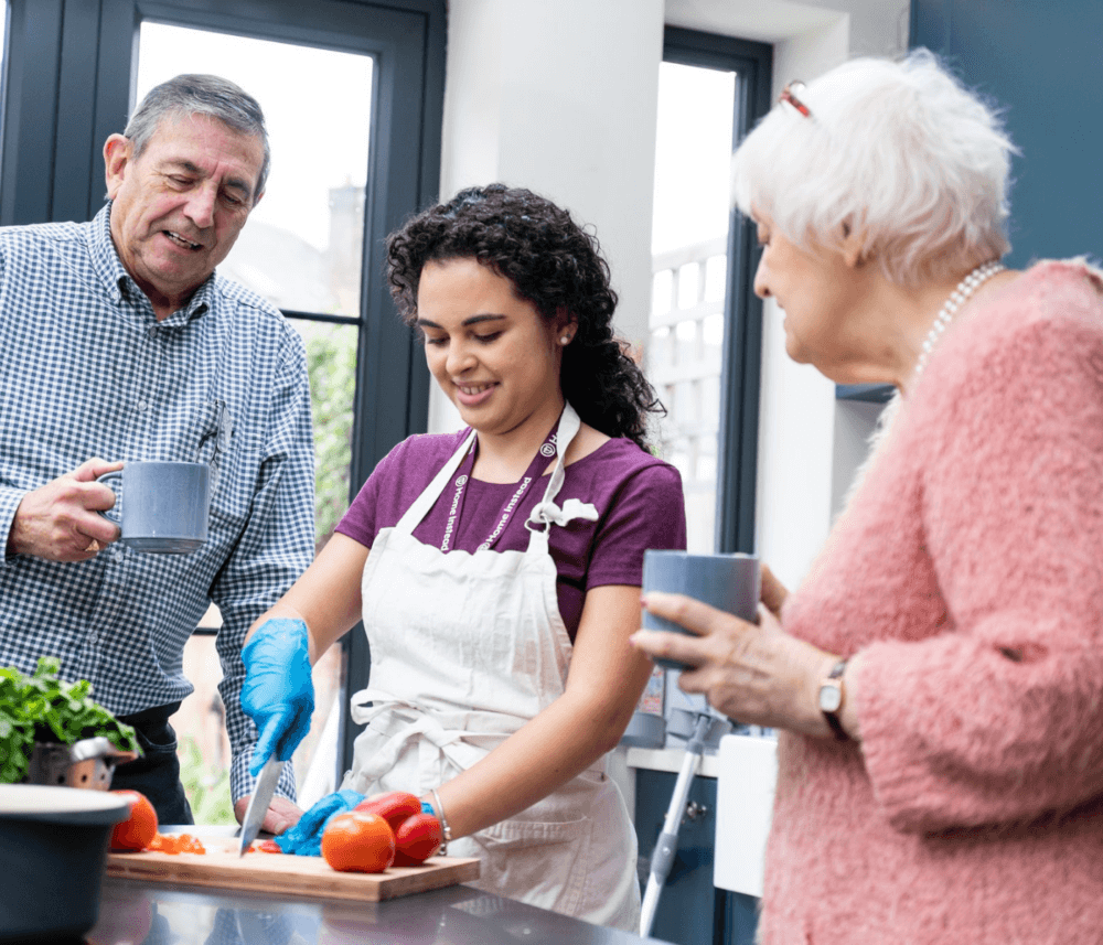 A person cuts vegetables while two others, each holding a mug, watch and smile in a bright kitchen. - Home Instead