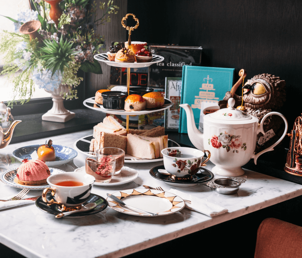 Elegant afternoon tea setup with a tiered tray of pastries, sandwiches, teapot, cups, and various decor on a marble table. - Home Instead