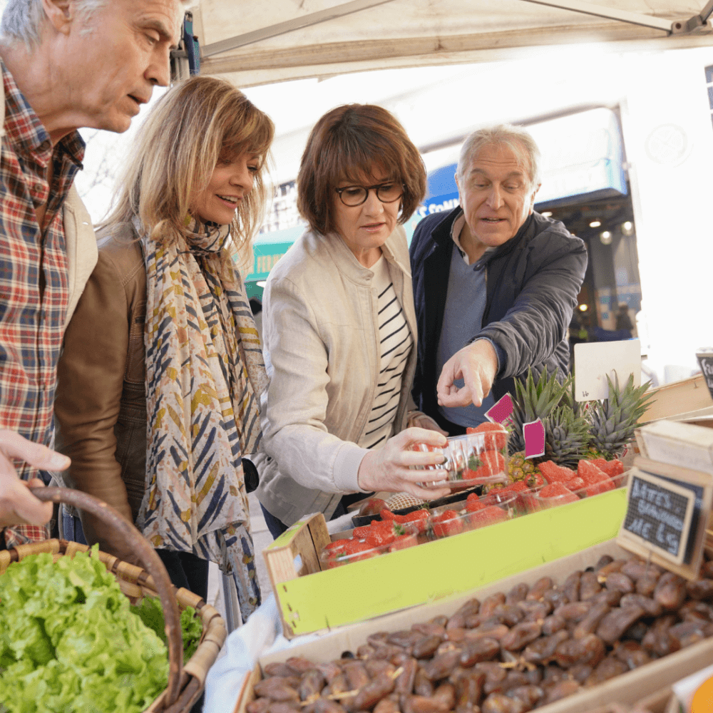 A group of people is shopping at an outdoor market stall, examining fresh produce and having a discussion. - Home Instead
