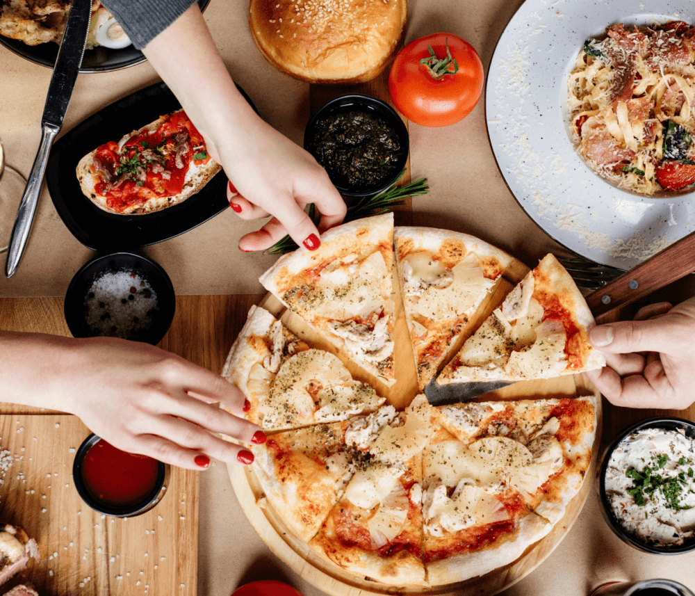 Hands reaching for slices of pizza on a table filled with various dishes and ingredients. - Home Instead
