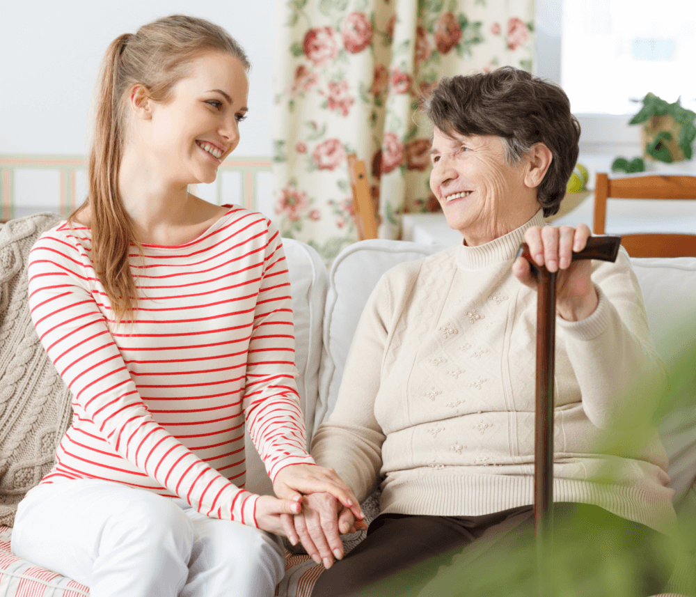 A young woman and an elderly woman sit on a sofa, smiling and holding hands. The elderly woman holds a cane. - Home Instead