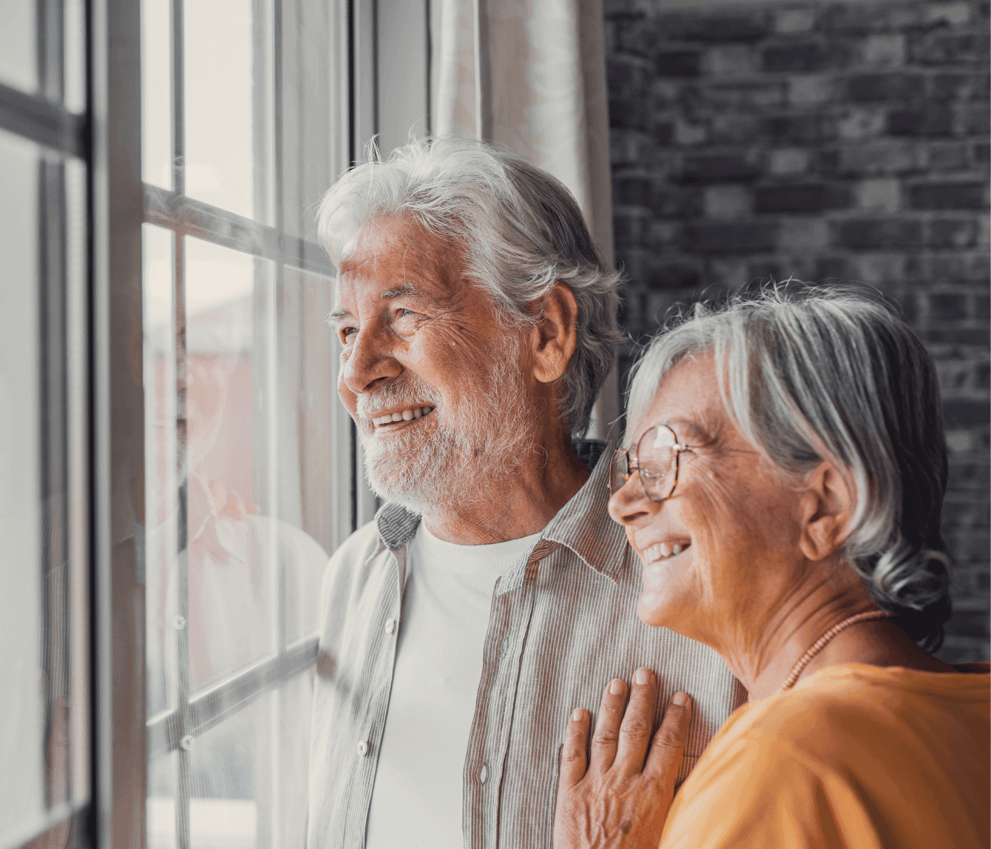 Elderly couple smiling and looking out of a window, sharing a moment of happiness indoors. - Home Instead