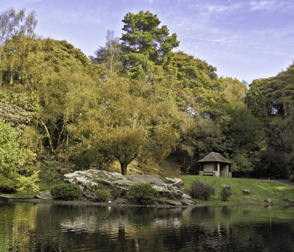 A small pavilion, lush trees, and a rocky area reflecting in a calm pond under a partly cloudy sky. - Home Instead