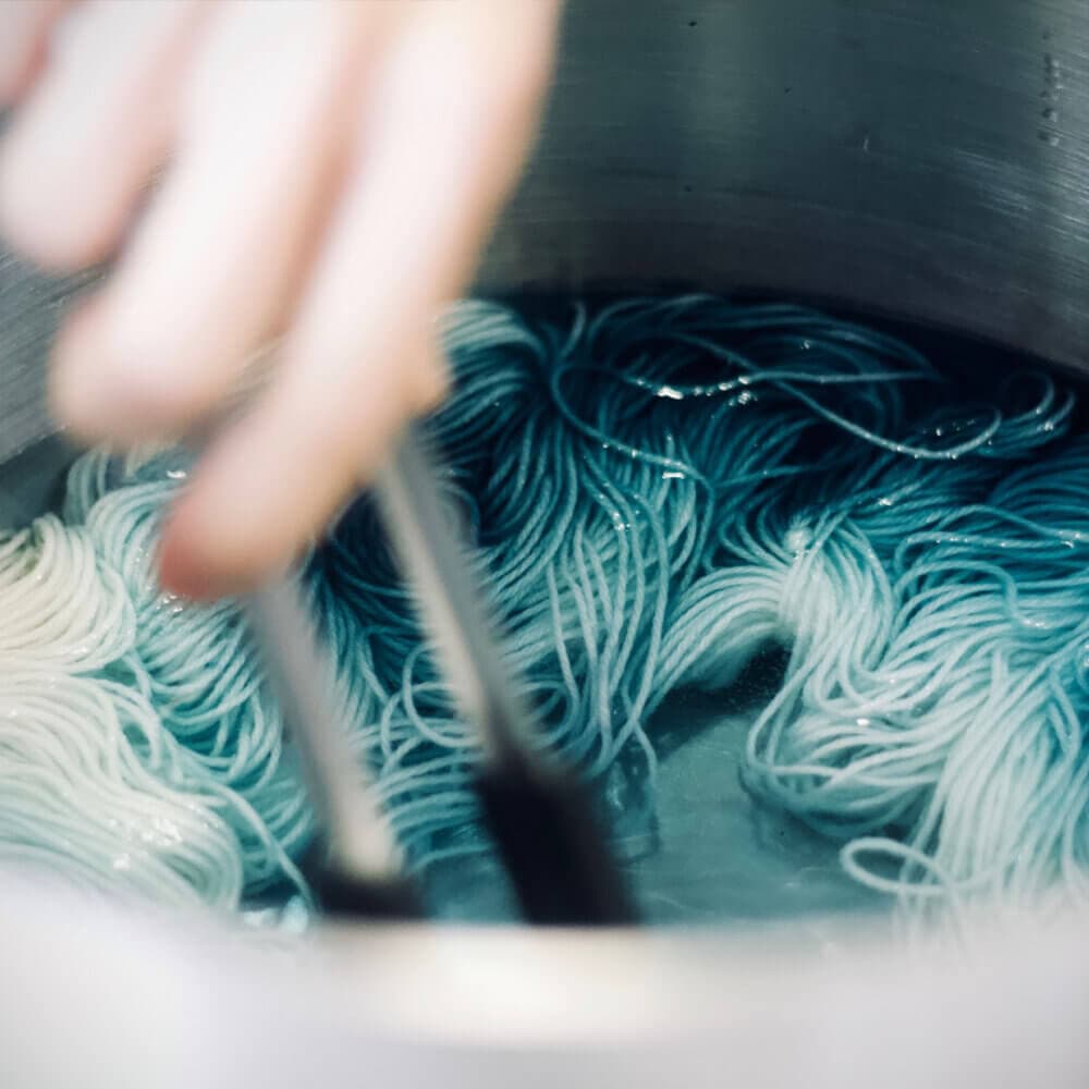 Hand using tongs to dye white yarn blue in a water-filled stainless steel pot. - Home Instead
