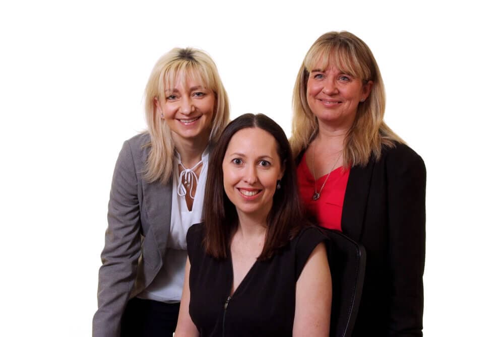 Three women are smiling at the camera; one is seated in front, with two standing on either side of her. - Home Instead