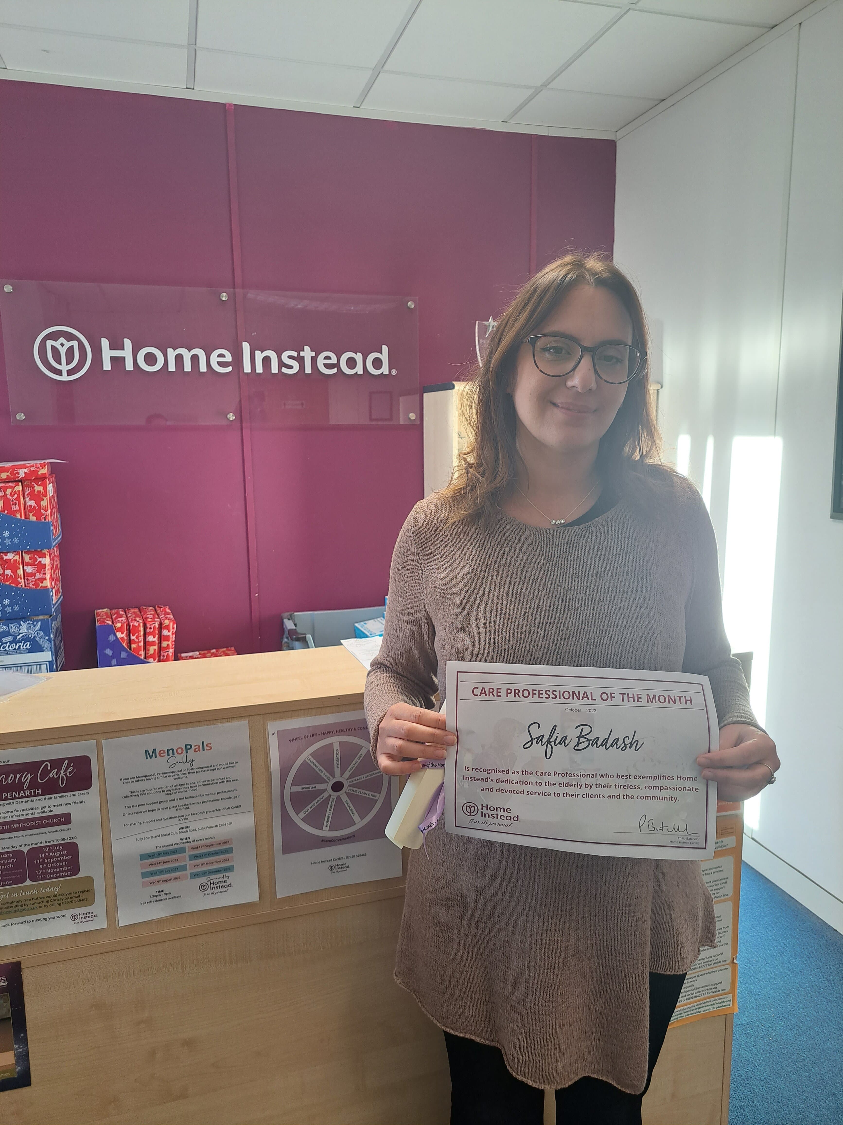 A person holding a "Care Professional of the Month" certificate in front of a Home Instead sign. - Home Instead