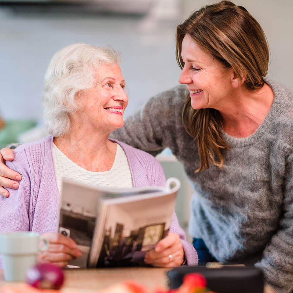 An elderly woman reading a magazine and smiling at a younger woman who is leaning over her shoulder and smiling back. - Home Instead