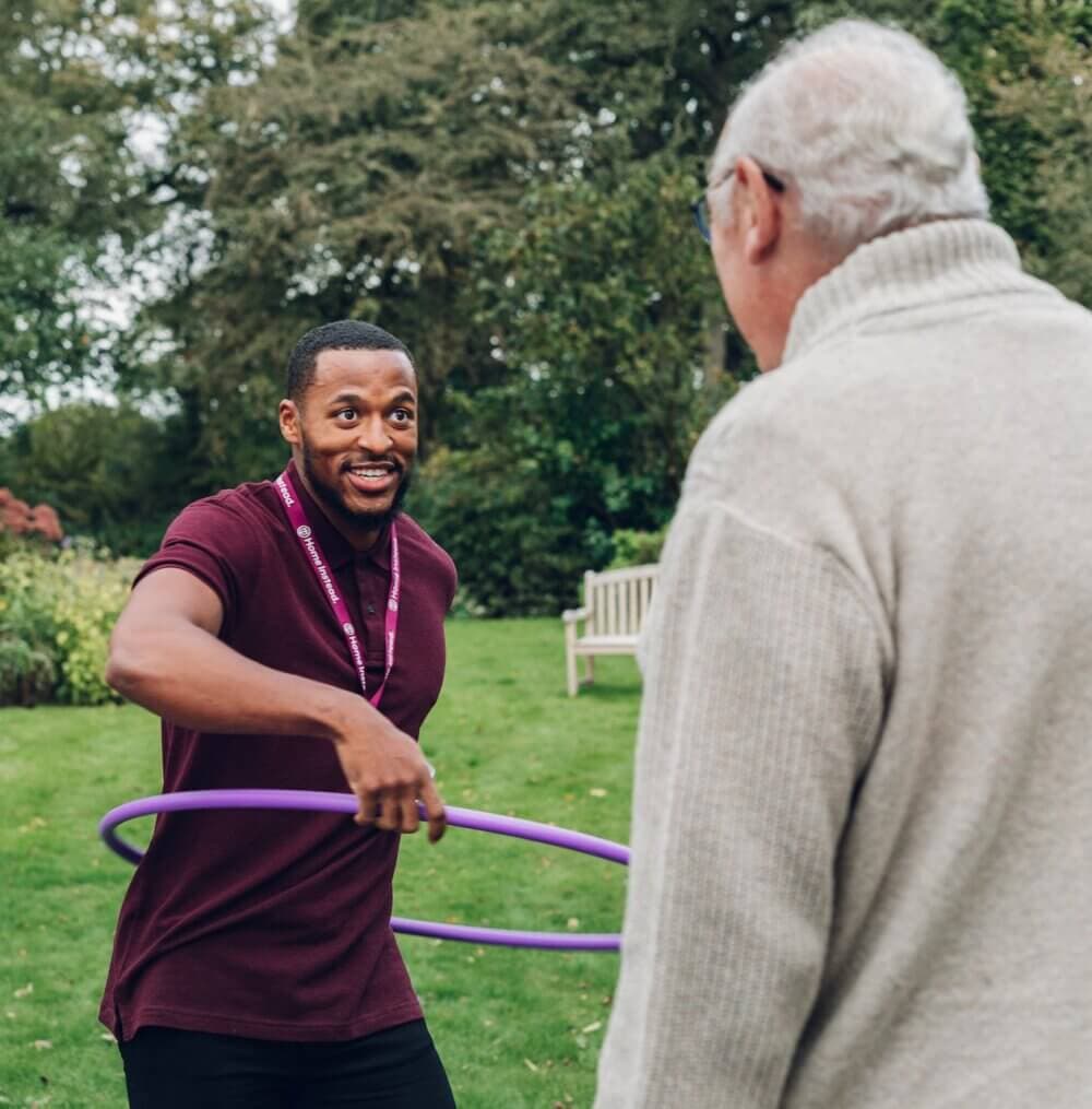 A man with a purple hula hoop smiles and interacts with an older man outdoors in a garden setting. - Home Instead