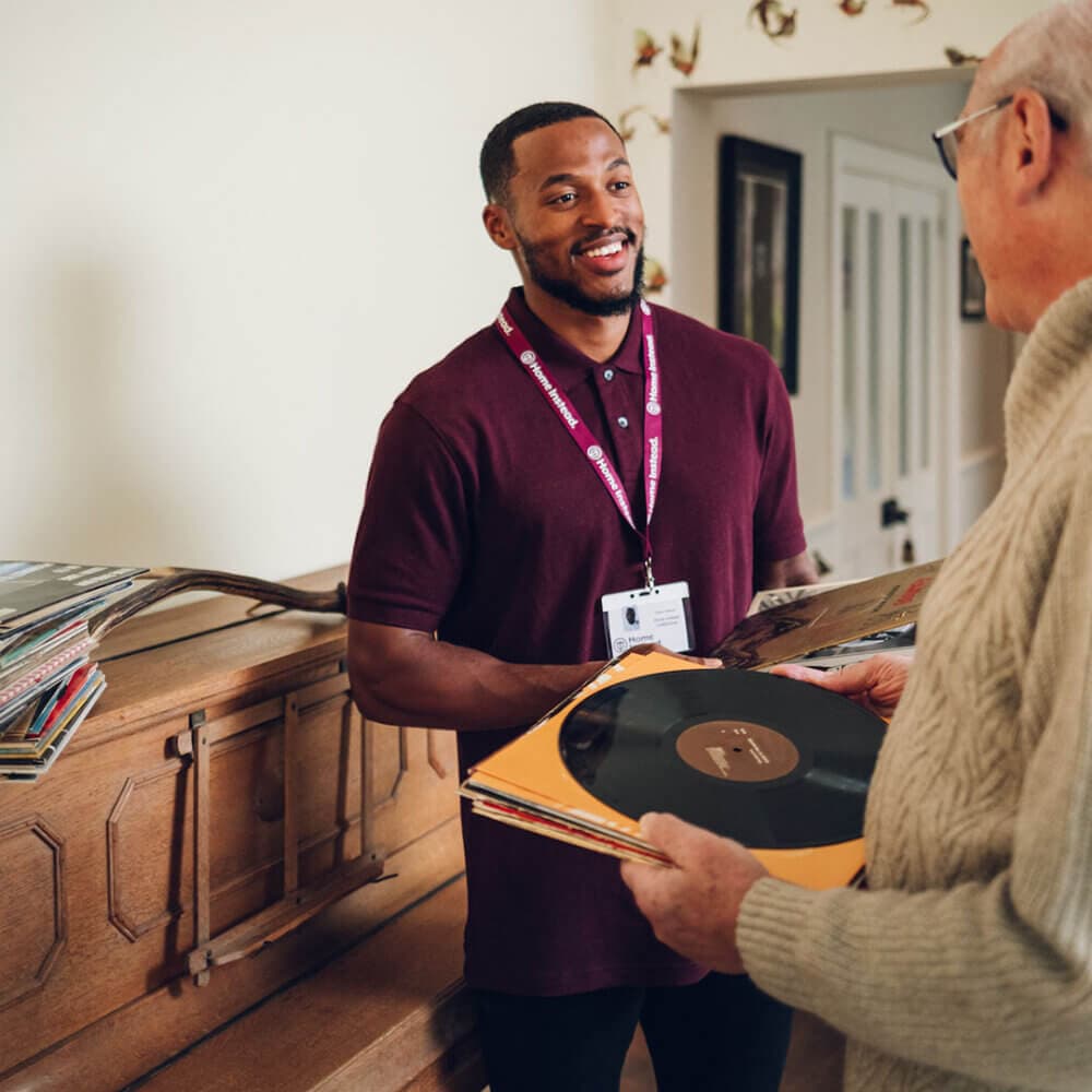 Man in burgundy shirt helps an elderly man holding vinyl records in a living room. - Home Instead