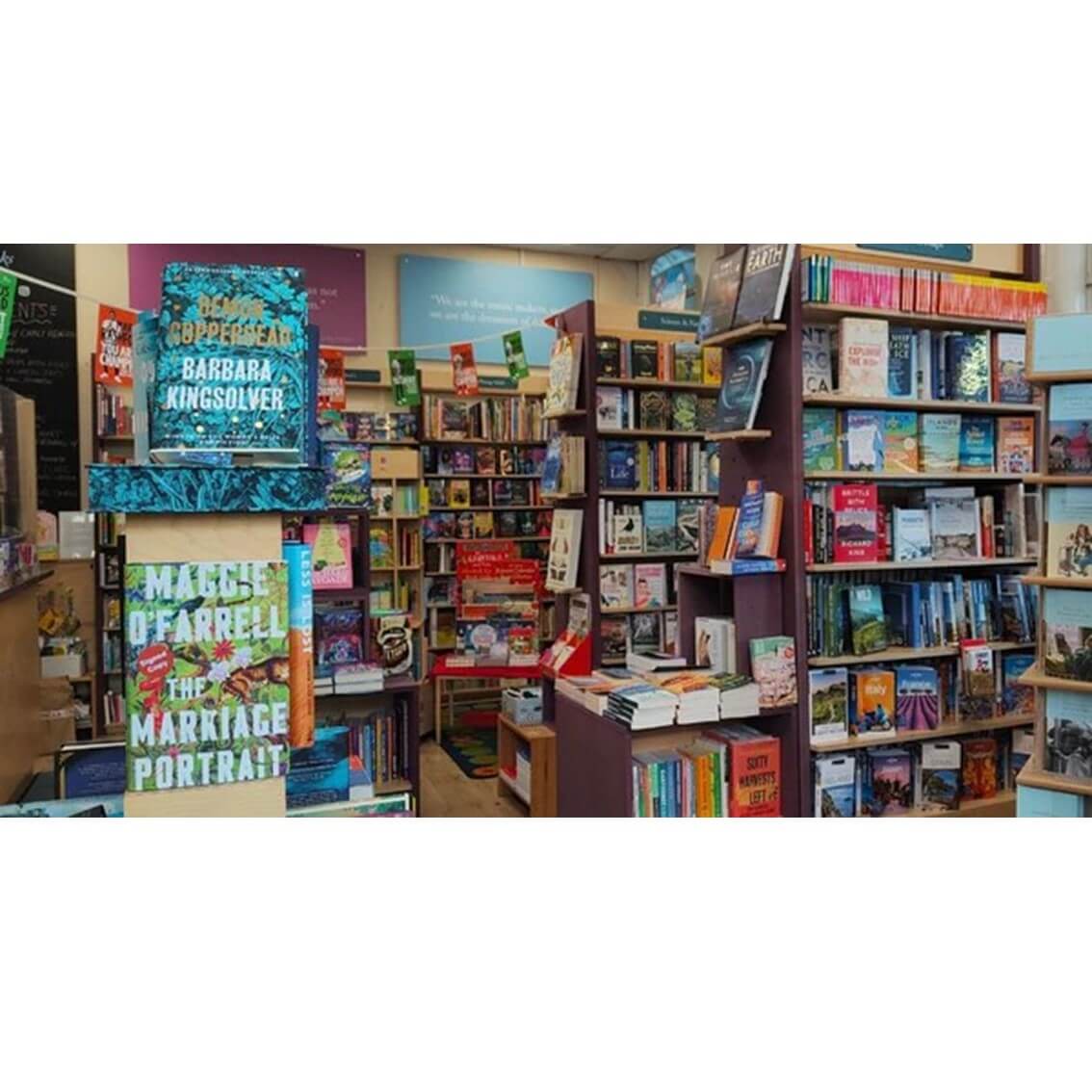 A bustling bookstore with shelves full of books, including prominent displays for "Demon Copperhead" and "The Marriage Portrait. - Home Instead