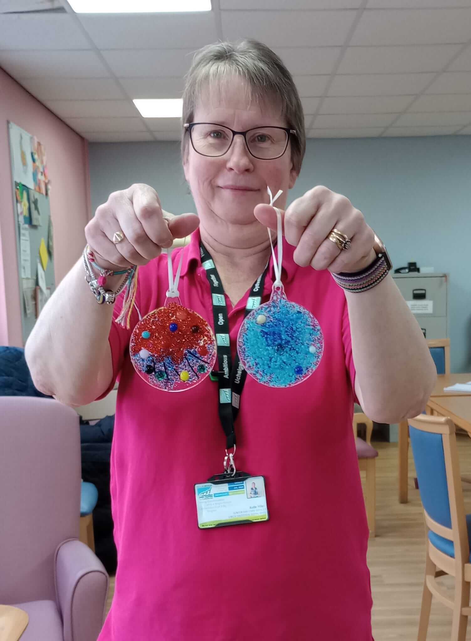 A person wearing glasses and a pink shirt holds up two round, colorful ornaments in a room with tables and chairs. - Home Instead