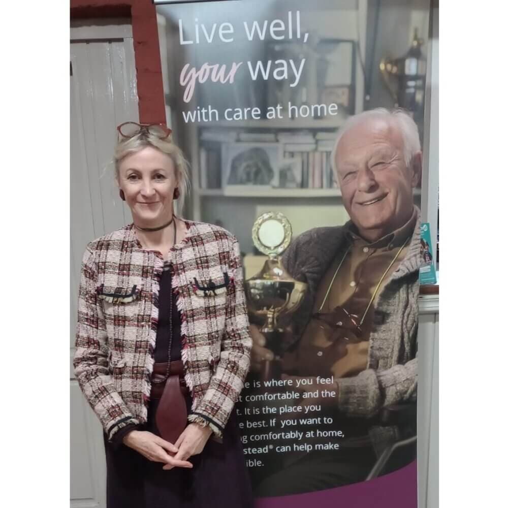 A woman in a patterned jacket stands in front of a banner featuring an elderly man and the text "Live well, your way. - Home Instead