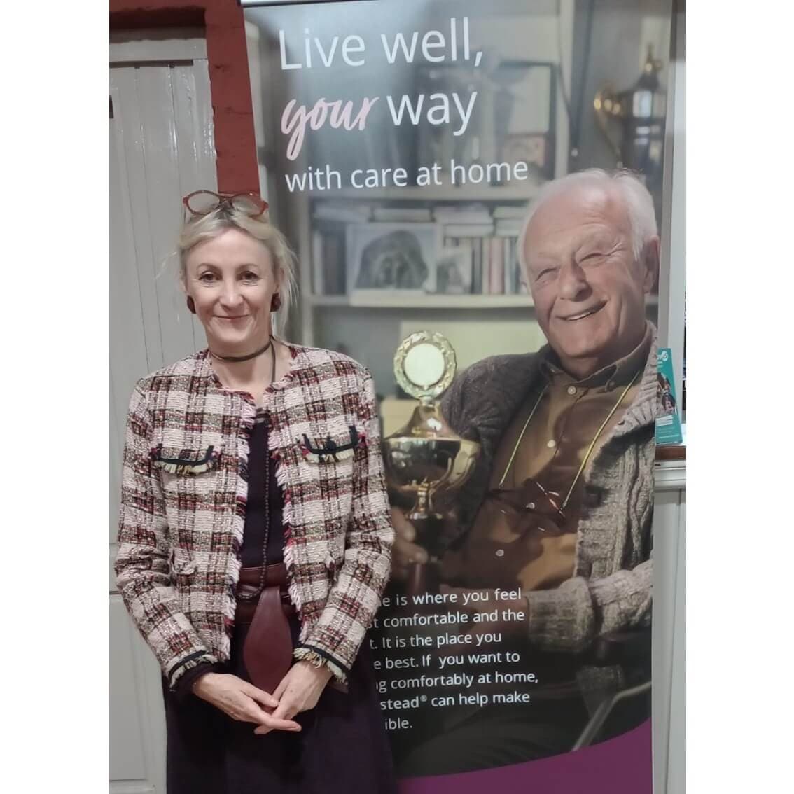 A woman in a patterned jacket stands in front of a banner featuring an elderly man and the text "Live well, your way. - Home Instead
