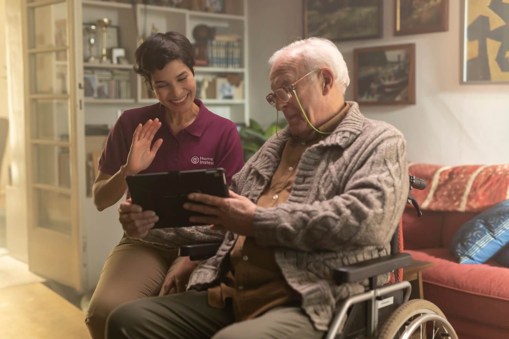 A smiling Care Professional waves while an elderly client in a wheelchair looks at a tablet in a cosy living room. - Home Instead Bournemouth & Christchurch