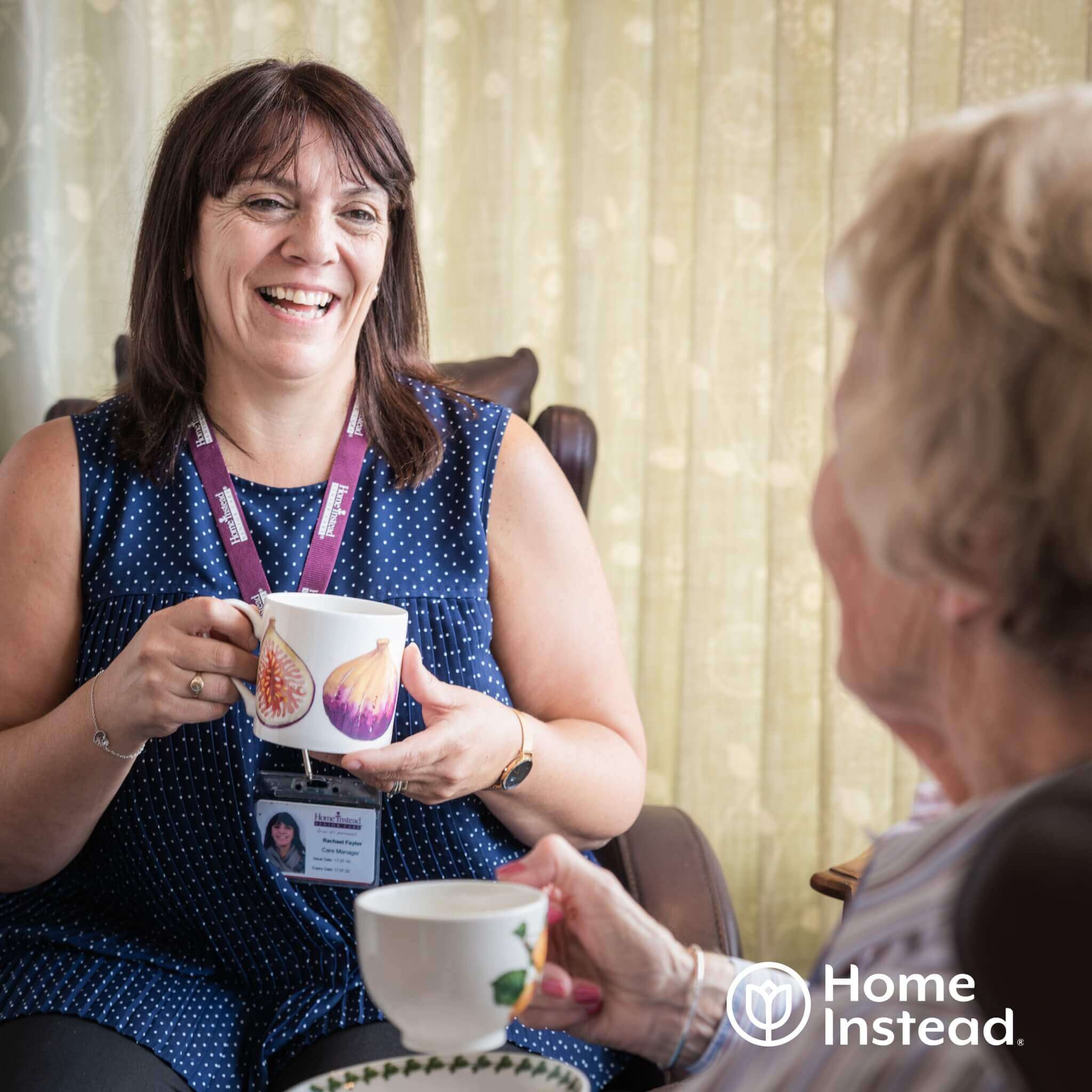 A caregiver and elderly person enjoy tea together, smiling. The image has the "Home Instead" logo in the bottom right. - Home Instead