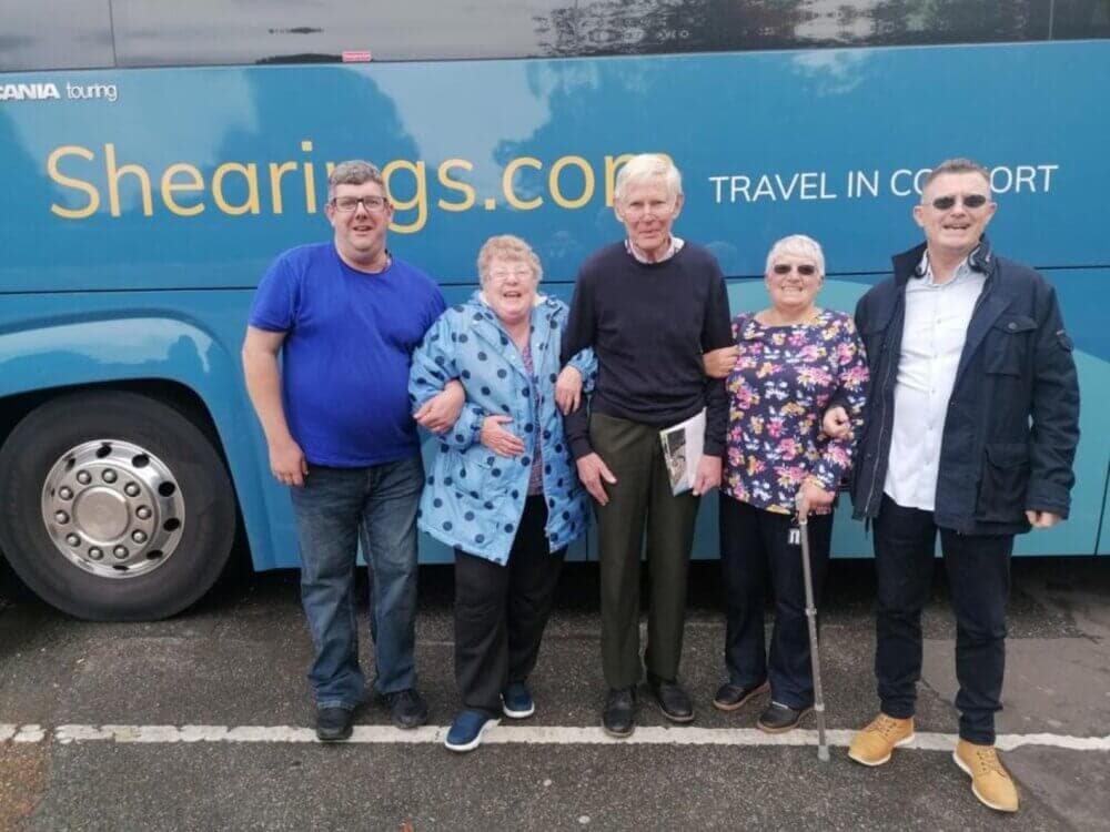 Five people are standing in front of a blue Shearings coach, smiling and posing for the photo. - Home Instead
