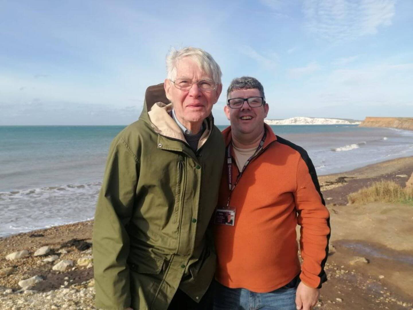 Two men standing on a rocky beach; the sea and cliffs can be seen in the background. - Home Instead