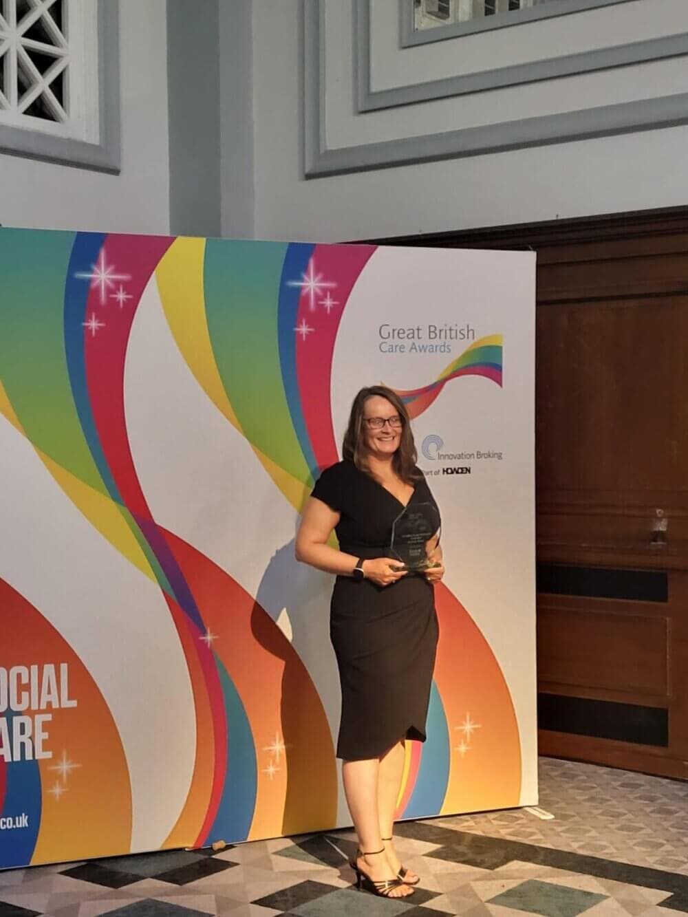 A woman in a black dress holding an award, standing in front of a colorful "Great British Care Awards" banner. - Home Instead
