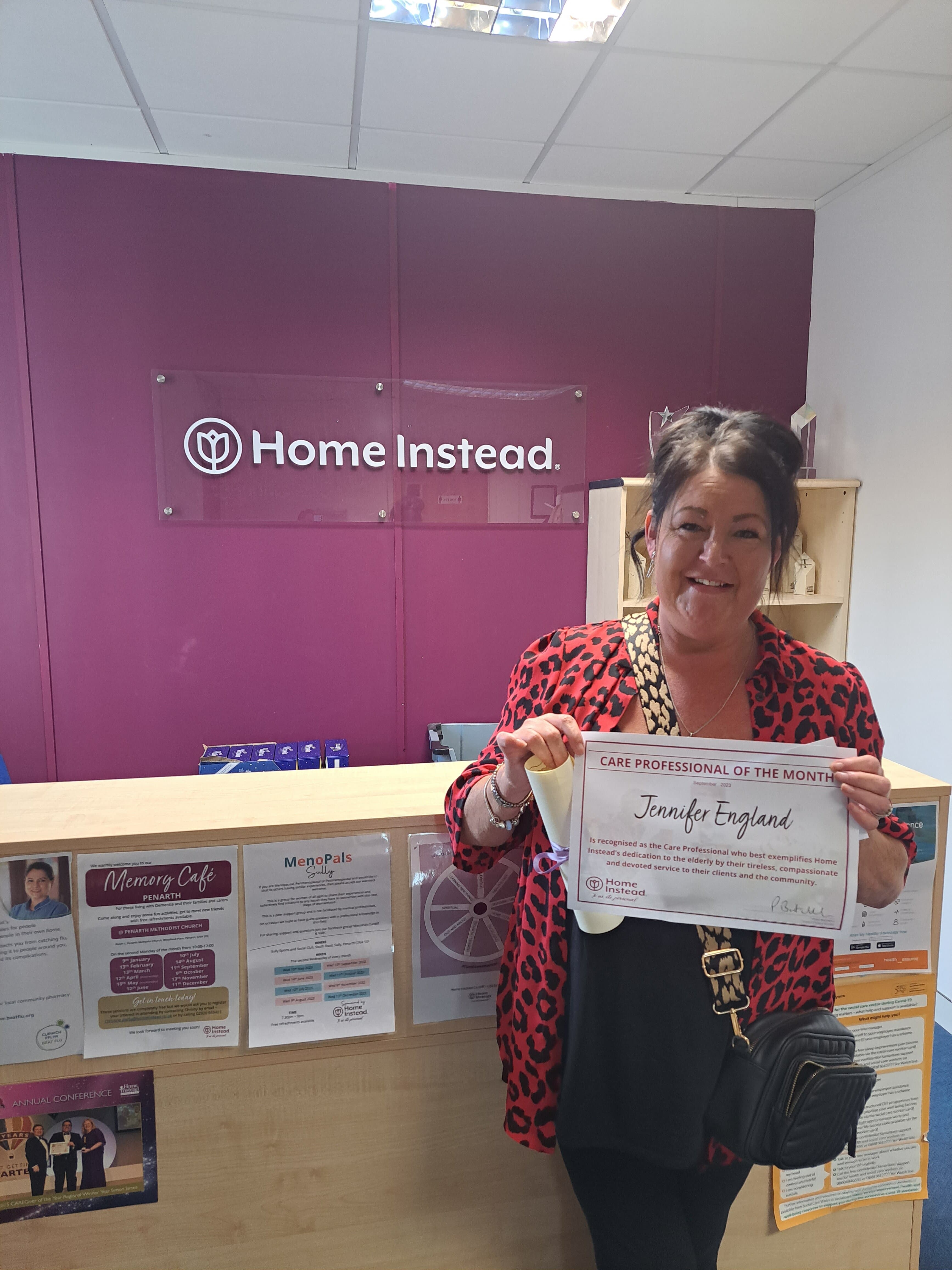 A woman stands smiling at a reception desk, holding a "Care Professional of the Month" certificate. - Home Instead