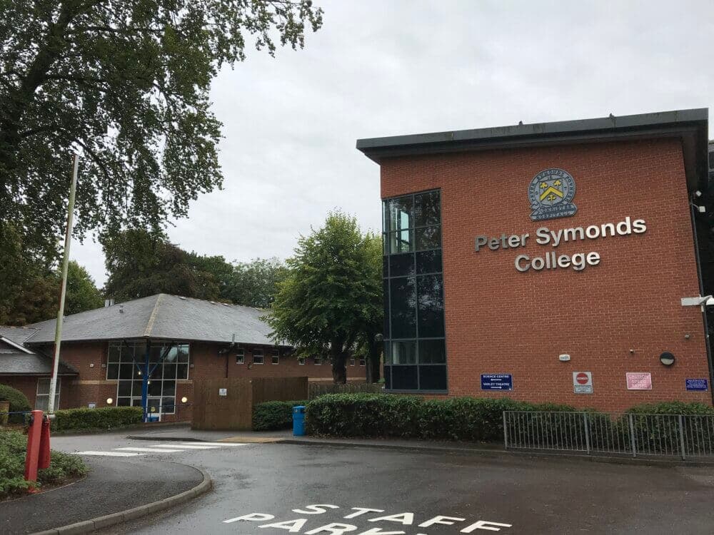 Red-brick building with "Peter Symonds College" sign, trees, and staff parking area. Cloudy sky in the background. - Home Instead
