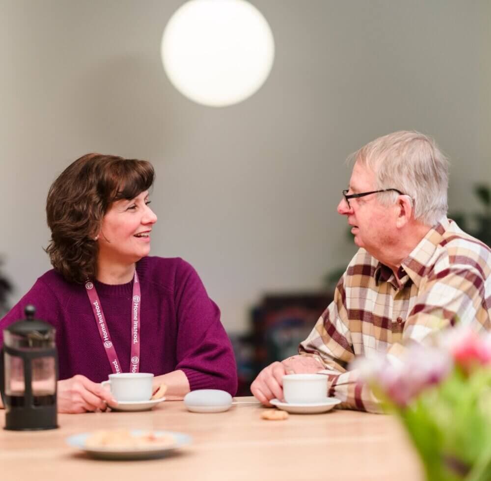 Two people sitting at a table, smiling and talking, with coffee cups and a smart speaker in front of them. - Home Instead