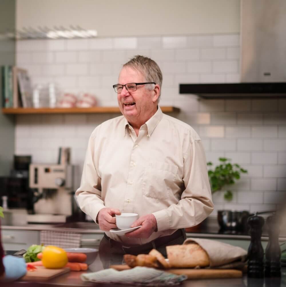 Elderly man smiling and holding a cup in a kitchen with vegetables and bread on the counter. - Home Instead
