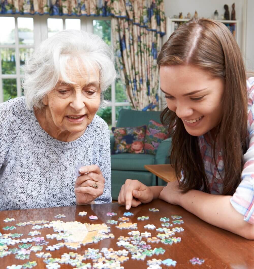 An elderly woman and a young woman work together on a jigsaw puzzle at a table, both smiling and engaged. - Home Instead