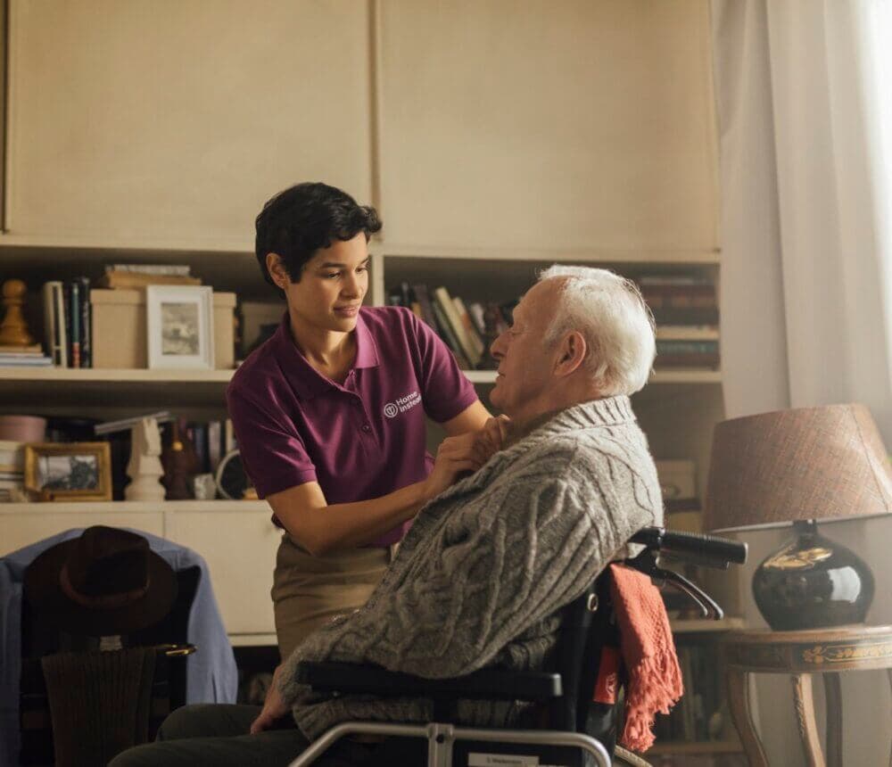 A caregiver in a purple polo shirt assists an elderly person in a wheelchair in a cozy, book-filled room. - Home Instead